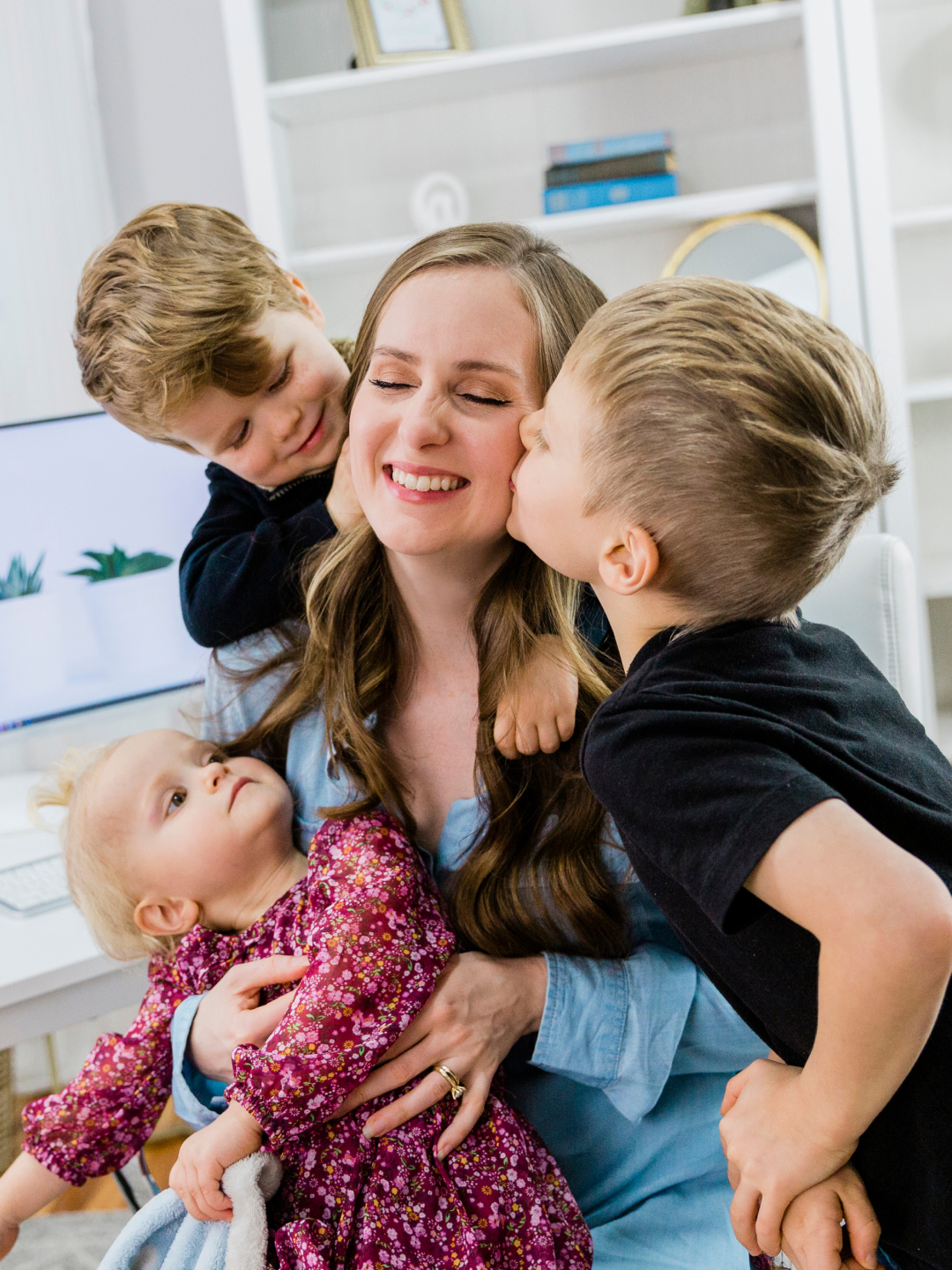 Gillian smiling in her home office as she holds her young daughter while her two sons hug and kiss her.